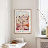 a framed poster of a colorful floral scene hanging on a wall, with a white table and a vase of flowers in the foreground.