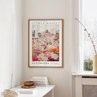 a framed poster of a colorful floral scene hanging on a wall, with a white table and a vase of flowers in the foreground.