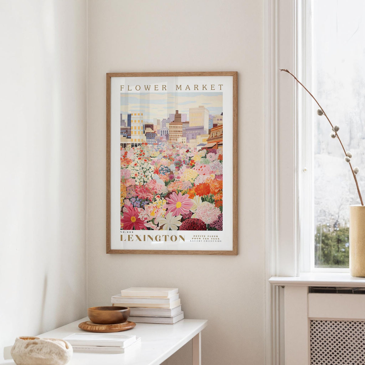 a framed poster of a colorful floral scene hanging on a wall, with a white table and a vase of flowers in the foreground.