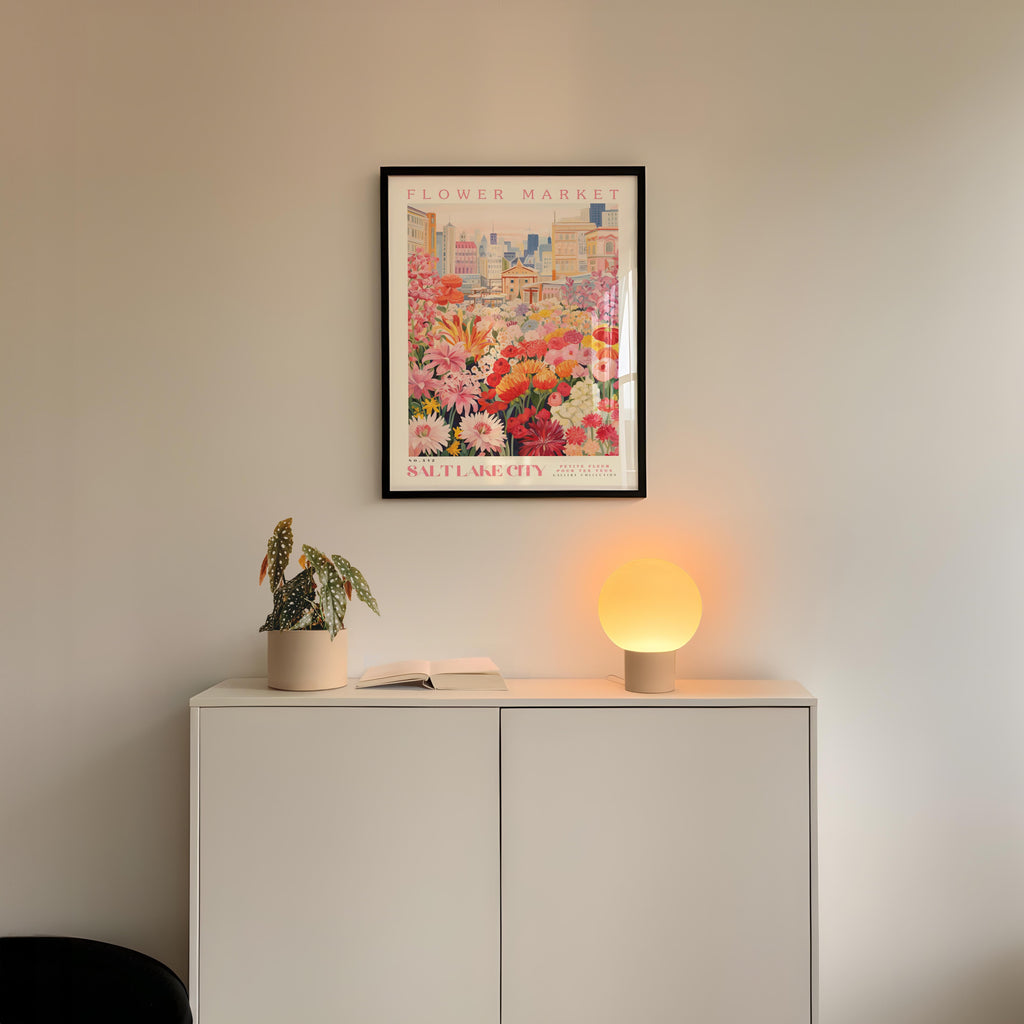 a white cabinet with a potted plant and a lamp on top, set against a wall with a framed poster of a cityscape above it.