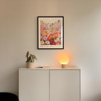 a white cabinet with a potted plant and a lamp on top, set against a wall with a framed poster of a cityscape above it.