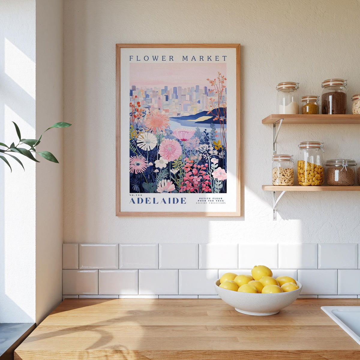 a framed poster of a flower market in Adelaide, Australia, hanging on a wall above a kitchen counter with a bowl of lemons and jars of food on the counter.