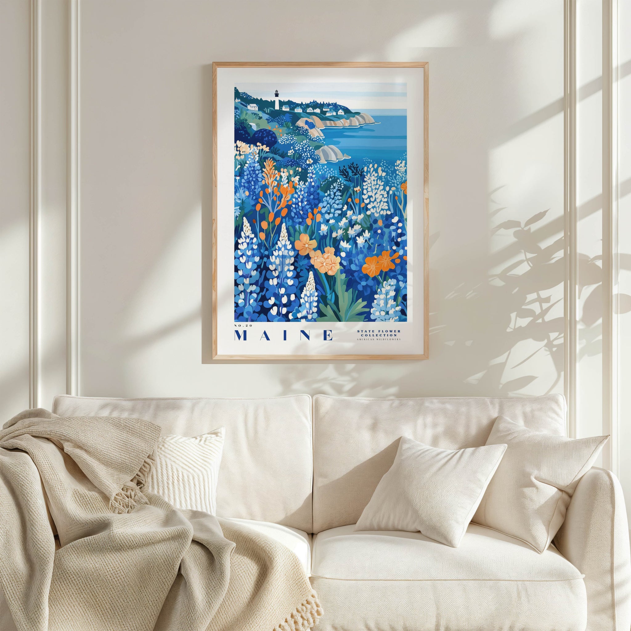 a framed poster of a coastal landscape with a lighthouse, hanging on a wall above a white couch with a blanket draped over it.