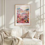 a framed poster of a colorful, floral-themed cityscape hanging on a wall above a white couch with pillows.