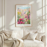 a framed poster of a colorful garden scene with flowers hanging on a wall above a white couch with pillows.