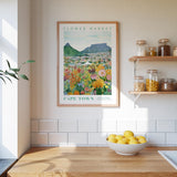 a framed poster of a colorful flower market in Cape Town, with a wooden shelf below it holding various jars and containers.