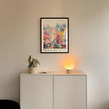 a white cabinet with a potted plant and a lamp on top, set against a wall with a framed poster of a colorful floral scene hanging above it.