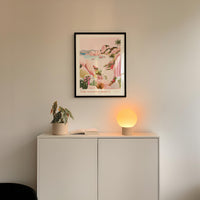 a white cabinet with a plant and a lamp on top, and a framed poster of a tropical landscape hanging on the wall above it.