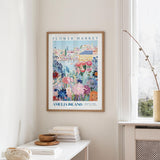 a framed poster of a colorful floral scene hanging on a wall, with a white table and a vase of flowers in the foreground.