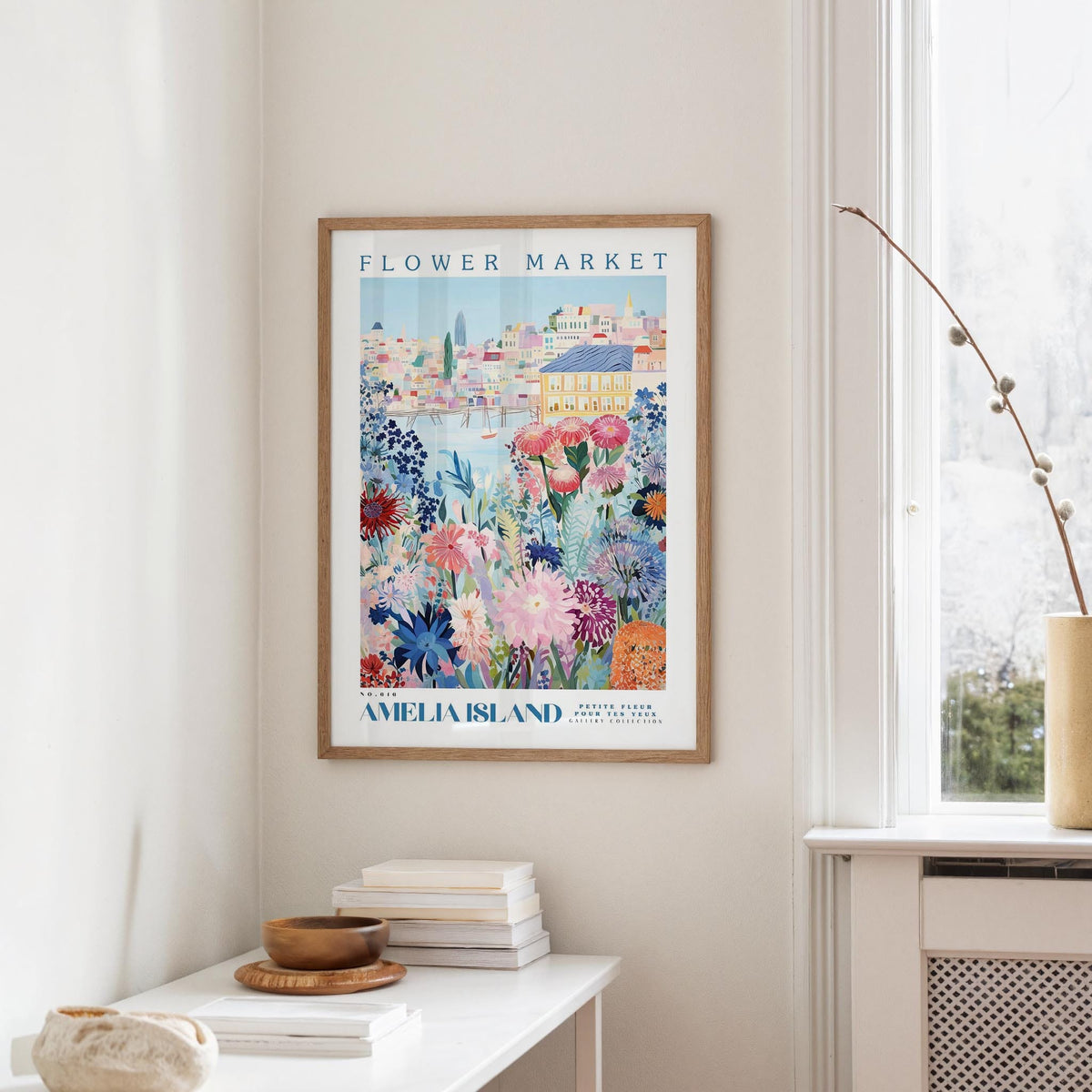 a framed poster of a colorful floral scene hanging on a wall, with a white table and a vase of flowers in the foreground.