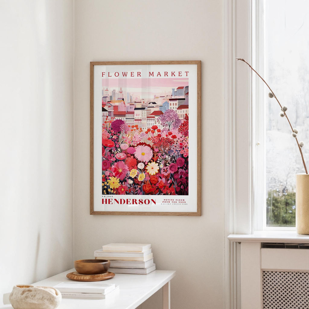 a framed poster of a colorful floral scene hanging on a wall, with a white table and a vase of flowers nearby.