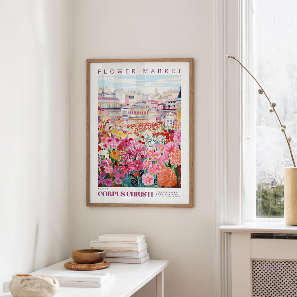 a framed poster of a colorful floral scene hanging on a wall, with a white table and a vase of flowers nearby.