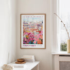 a framed poster of a colorful floral scene hanging on a wall, with a white table and a vase of flowers nearby.