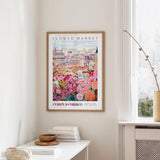 a framed poster of a colorful floral scene hanging on a wall, with a white table and a vase of flowers nearby.