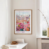 a framed poster of a colorful floral scene hanging on a wall, with a white table and a vase of flowers nearby.