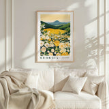 a framed poster of a field of flowers, with the word "GEORGIA" prominently displayed. The poster is hanging on a wall above a white couch, which is adorned with a blanket and pillows.