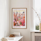 a framed poster of a cityscape with a flower market in the background, hanging on a wall in a room with a window and a small table with books and a vase.