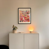 a white cabinet with a potted plant and a lamp on top, set against a white wall. Above the cabinet, there is a framed poster of a colorful cityscape.