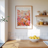 a kitchen scene with a framed poster of a colorful floral market hanging on the wall above a wooden counter. On the counter, there is a bowl of lemons and a plant. The kitchen has white tiled walls and a wooden shelf with various jars and containers.