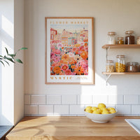 a kitchen scene with a framed poster of a colorful floral market hanging on the wall above a wooden counter. On the counter, there is a bowl of lemons and a plant. The kitchen has white tiled walls and a wooden shelf with various jars and containers.