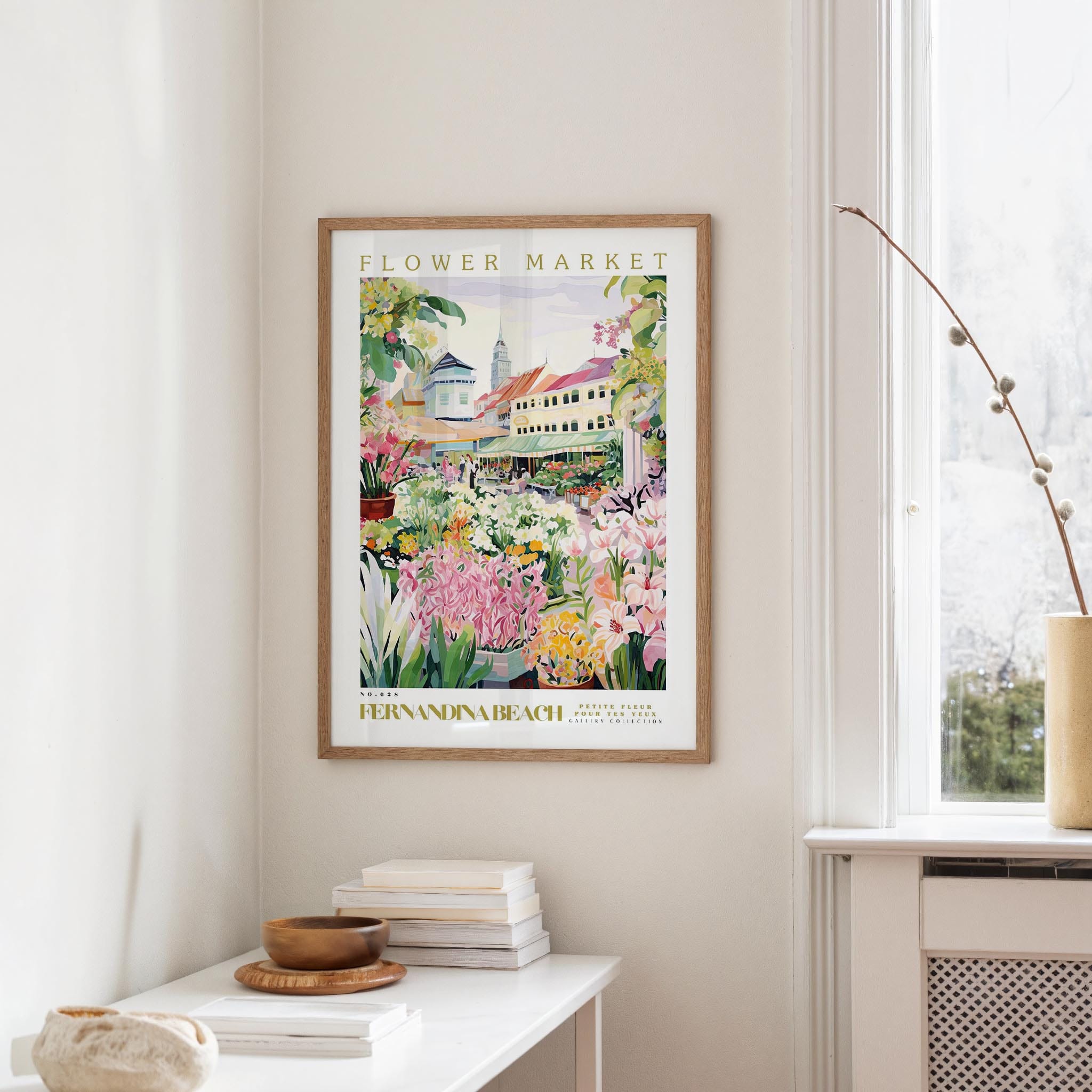 a framed poster of a colorful garden scene hanging on a wall, with a white table and a vase of flowers in the foreground.