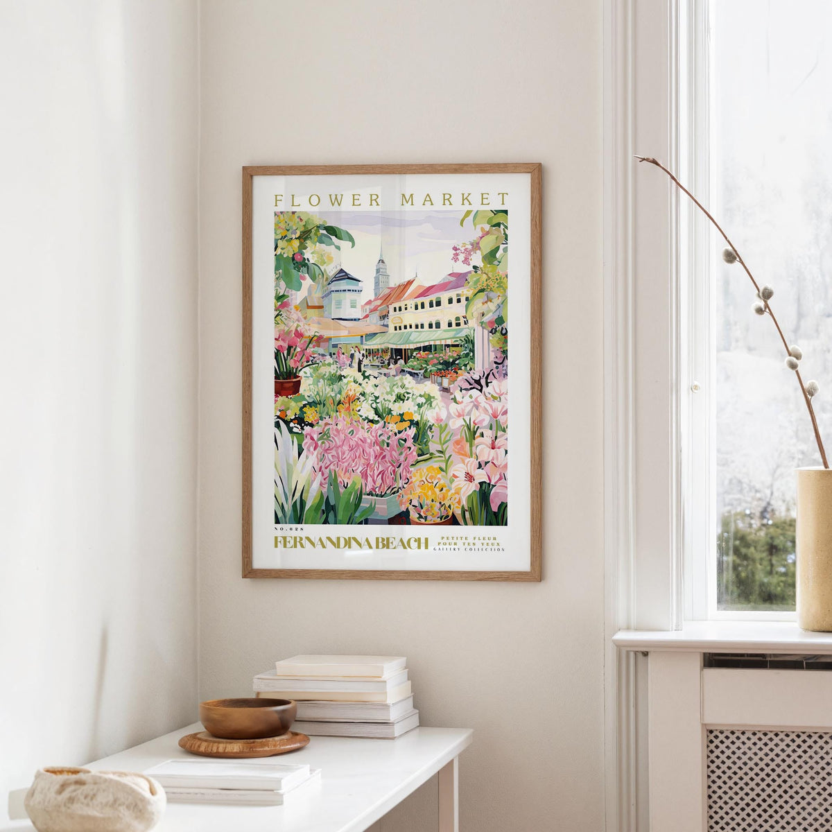 a framed poster of a colorful garden scene hanging on a wall, with a white table and a vase of flowers in the foreground.