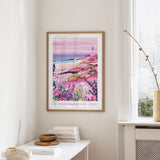a framed poster of a coastal landscape with a lighthouse, hanging on a wall in a room with a white table and a window.