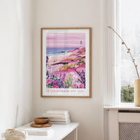 a framed poster of a coastal landscape with a lighthouse, hanging on a wall in a room with a white table and a window.