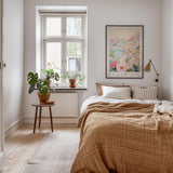 a cozy bedroom with a large bed, a nightstand with a potted plant, and a framed poster of a cityscape on the wall.