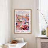 a framed poster of a colorful floral scene hanging on a wall, with a window and a small table with books and a vase in the foreground.