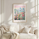 a framed poster of a cityscape with a field of flowers in the foreground, hanging on a wall above a white couch with a blanket draped over it.