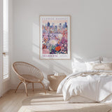 a bedroom with a large poster of the Melbourne Flower Market hanging on the wall. The poster depicts a vibrant cityscape with a variety of flowers and plants. The room has a white bed, a woven chair, and a small side table. The overall color scheme of the room is predominantly white, with the poster adding a pop of color and interest to the space.