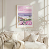 a framed poster of a beach scene with a pink and purple sky, palm trees, and a body of water. The poster is hanging on a wall above a white couch with a blanket draped over it.