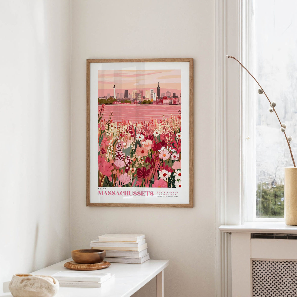 a framed poster of a cityscape with a field of flowers in the foreground, hanging on a wall above a white table with a stack of books and a small vase.