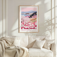a framed poster of a scenic landscape with a field of blooming flowers, including pink and white flowers, against a backdrop of mountains and a lake. The poster is hung on a white wall above a white couch, which has a blanket draped over it.
