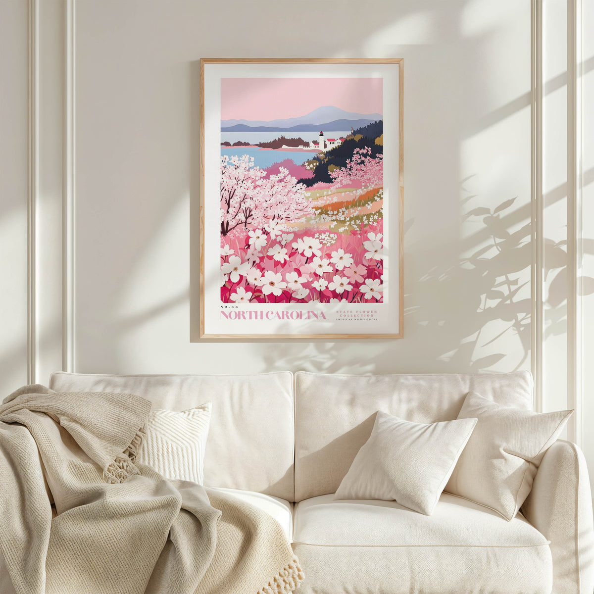 a framed poster of a scenic landscape with a field of blooming flowers, including pink and white flowers, against a backdrop of mountains and a lake. The poster is hung on a white wall above a white couch, which has a blanket draped over it.