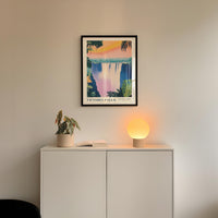 a white cabinet with a plant and a lamp on top, and a framed poster of a waterfall hanging on the wall above it.