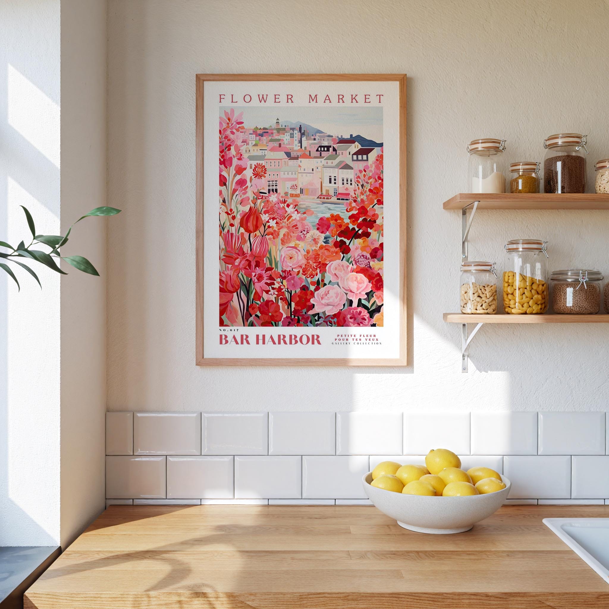 a kitchen scene with a framed poster of a flower market hanging on the wall above a wooden counter. On the counter, there is a bowl of lemons and a plant. The kitchen has white tile backsplash and shelves with various jars and containers.