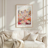 a framed poster of a flower market in a city, hanging on a wall above a white couch with a blanket draped over it.