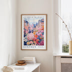 a framed poster of a colorful floral scene hanging on a wall, with a white table and a vase of flowers in the foreground.
