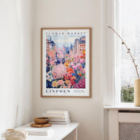 a framed poster of a colorful floral scene hanging on a wall, with a white table and a vase of flowers in the foreground.