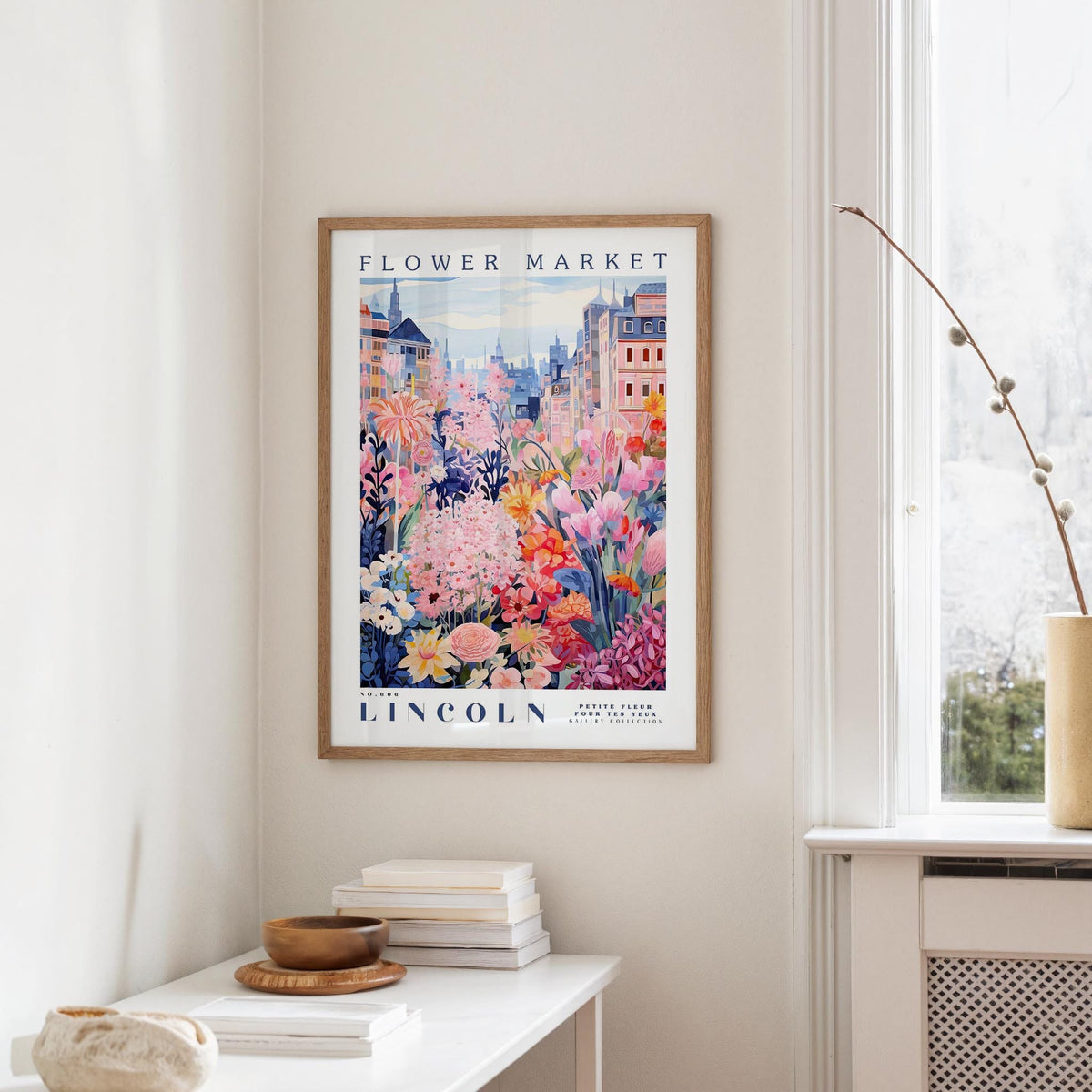 a framed poster of a colorful floral scene hanging on a wall, with a white table and a vase of flowers in the foreground.