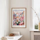 a framed poster of a colorful floral scene hanging on a wall, with a white table and a vase of flowers nearby.