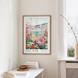 a framed poster of a colorful garden scene hanging on a wall, with a white table and a vase of flowers in the foreground.