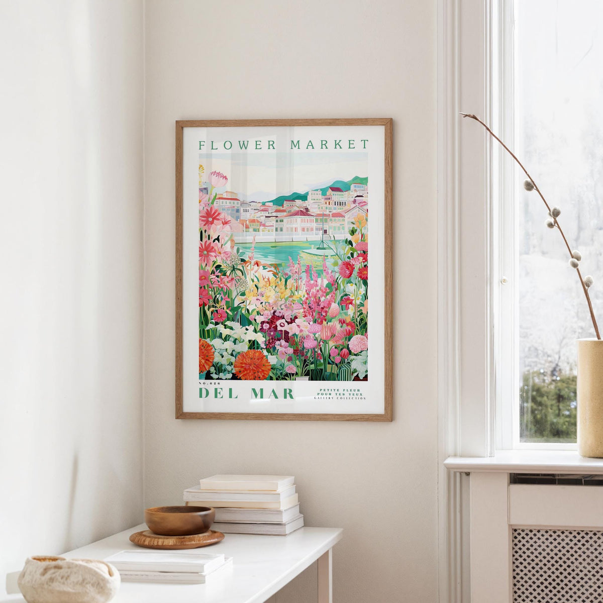 a framed poster of a colorful garden scene hanging on a wall, with a white table and a vase of flowers in the foreground.