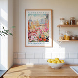 a kitchen scene with a framed poster of a colorful flower market in Des Moines, Iowa, hanging on the wall above a wooden counter. The poster is framed in wood and has a white border. On the counter, there is a bowl of lemons and a plant. The kitchen has white tile walls and a wooden floor.