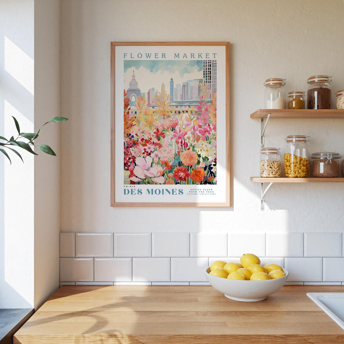 a kitchen scene with a framed poster of a colorful flower market in Des Moines, Iowa, hanging on the wall above a wooden counter. The poster is framed in wood and has a white border. On the counter, there is a bowl of lemons and a plant. The kitchen has white tile walls and a wooden floor.