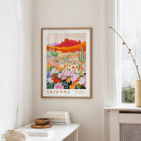 a framed poster of a desert landscape with cacti and flowers, hanging on a wall in a room with a white table and a window.