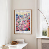 a framed poster of a cityscape with a flower market in the background, hanging on a wall in a room with a window and a table with books and a bowl on it.
