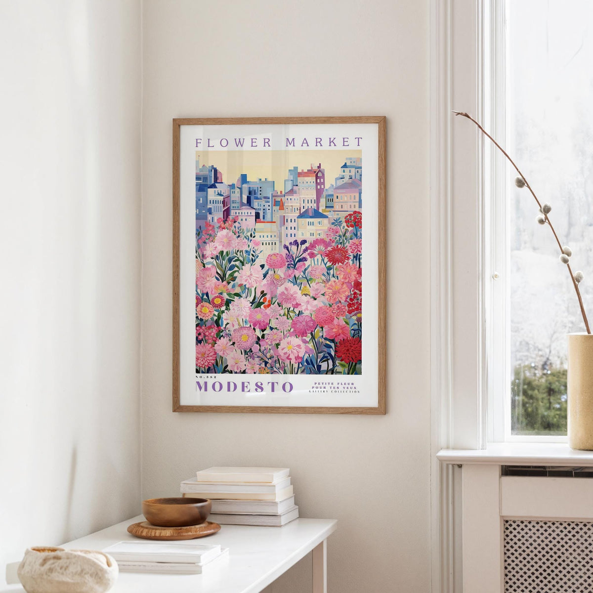 a framed poster of a cityscape with a flower market in the background, hanging on a wall in a room with a window and a table with books and a bowl on it.